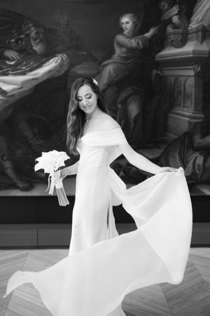 A bride in a flowing white long-sleeved gown twirls gracefully while holding a bouquet against an ornate dark background.