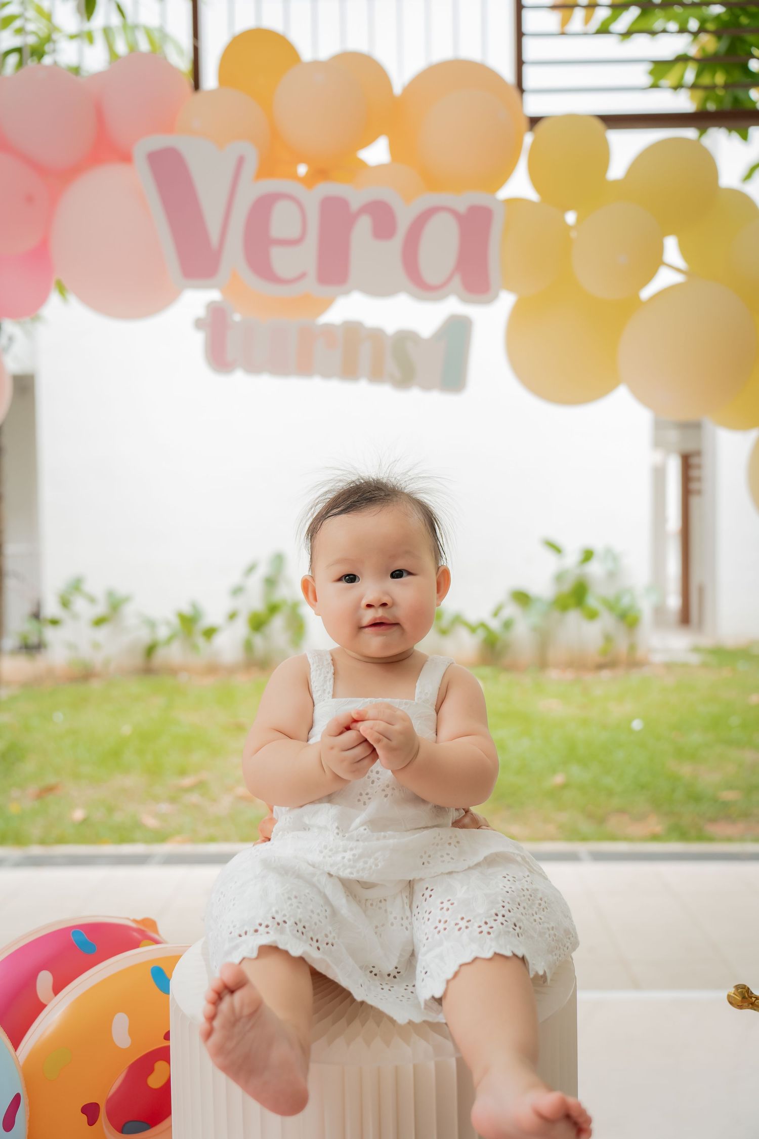 Baby in white dress sits under pastel balloon decorations at a party.
