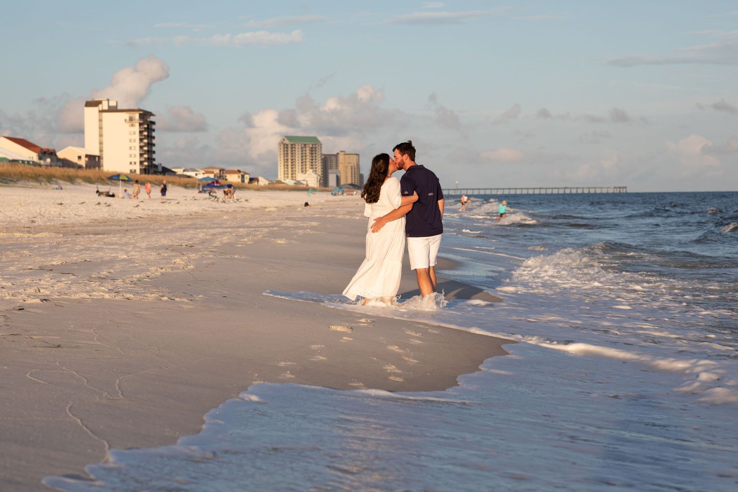 Coastal photography session on white sandy beach with waves crashing and buildings in background.