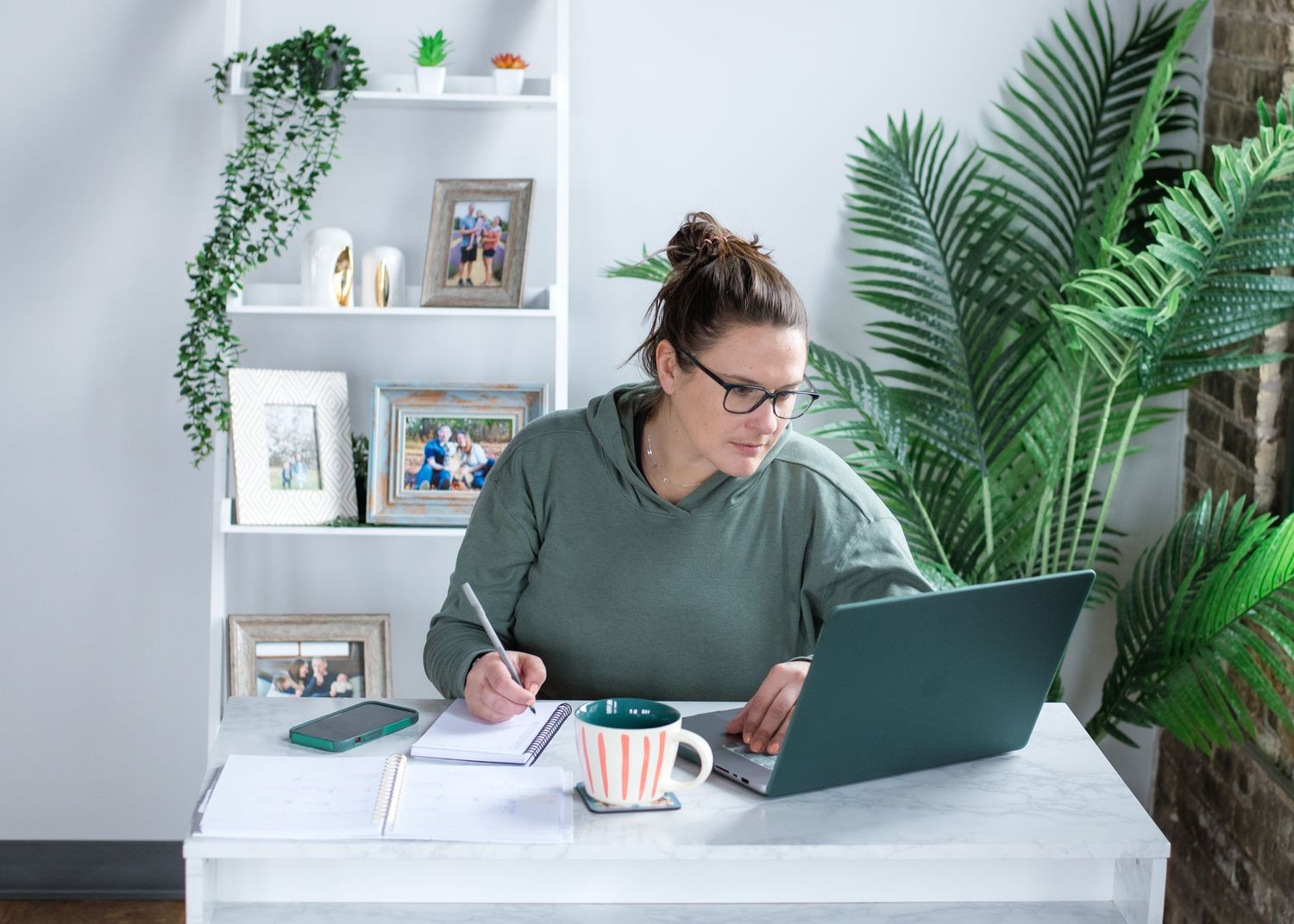 Person in green sweater working at desk with laptop surrounded by plants and modern decor.