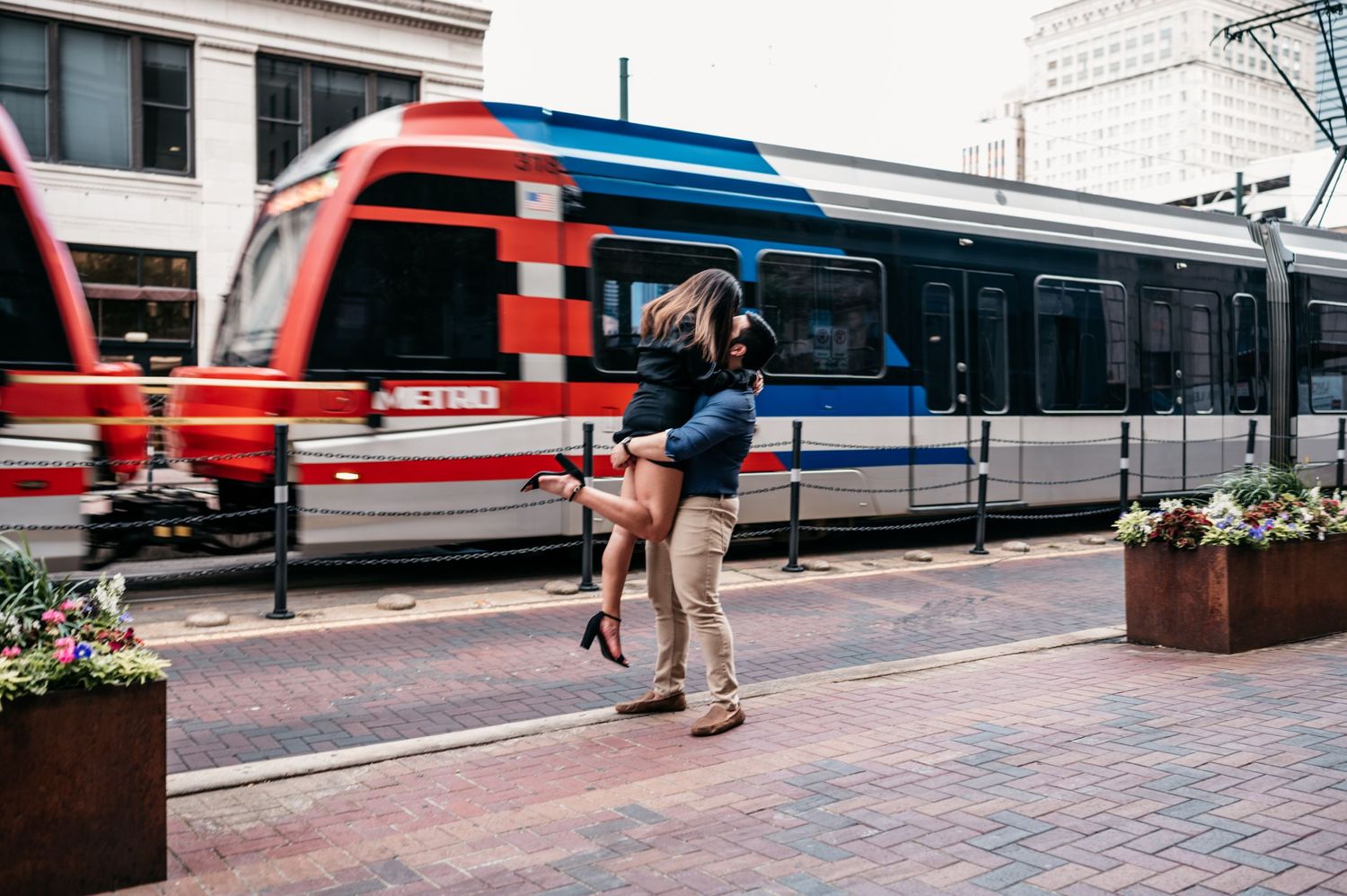 A couple embraces on city streets with light rail trains in the background.