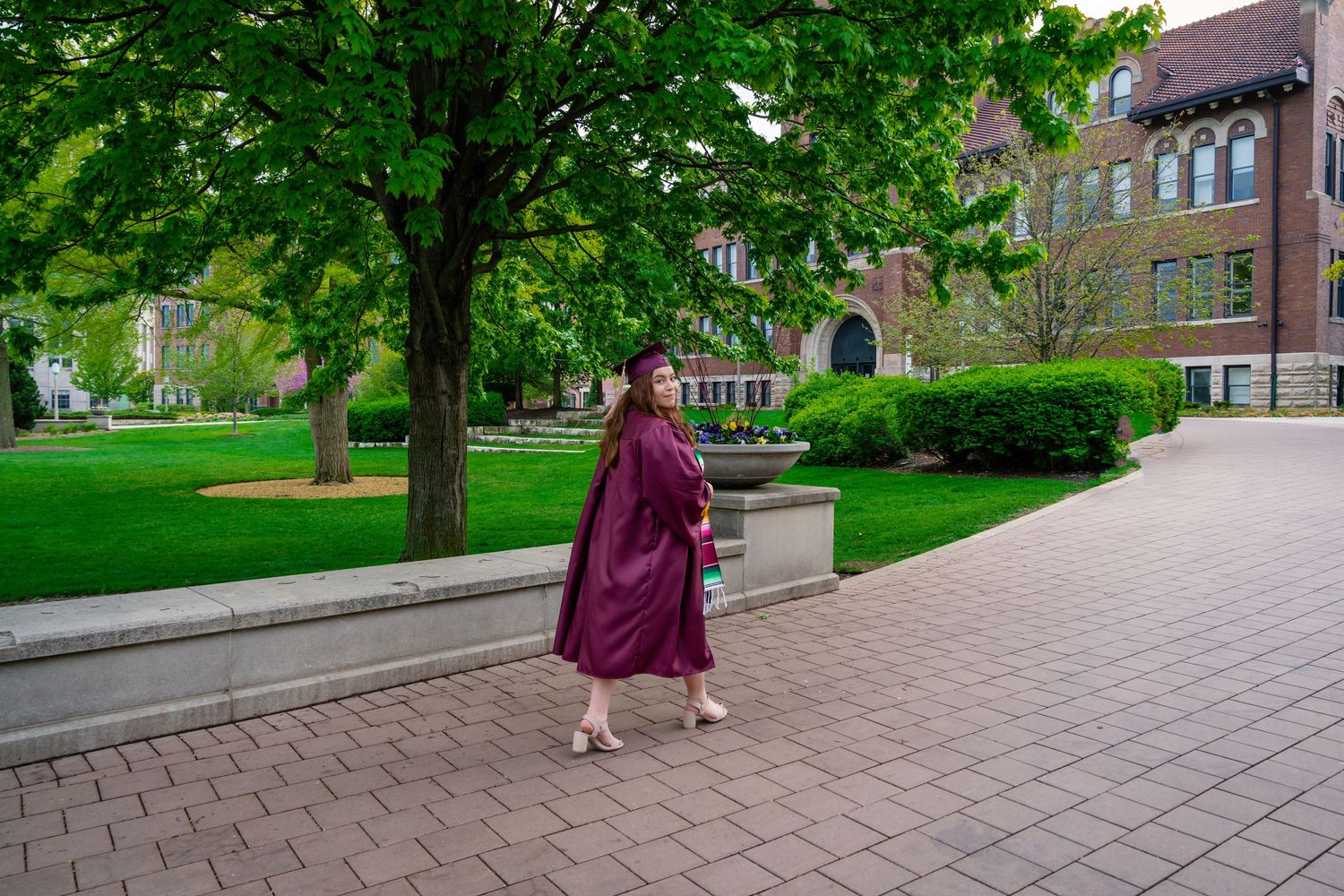 Person in burgundy sweater walking along brick pathway surrounded by lush green trees and buildings.
