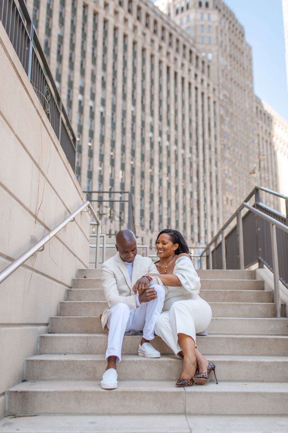 A stylish couple in all-white outfits sharing a moment on concrete steps near a city building.