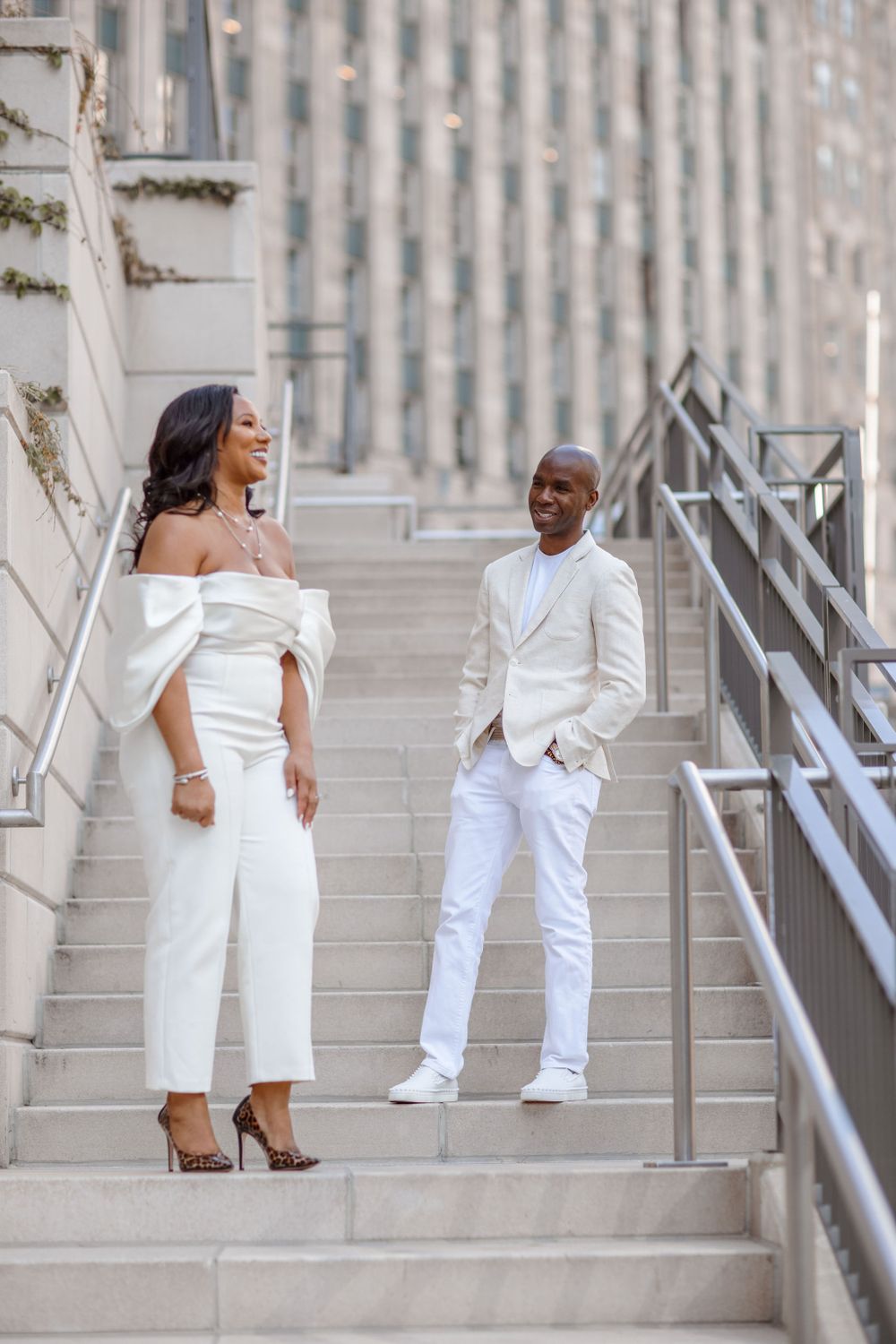 A stylish couple in white attire poses on stone steps outside an urban building.