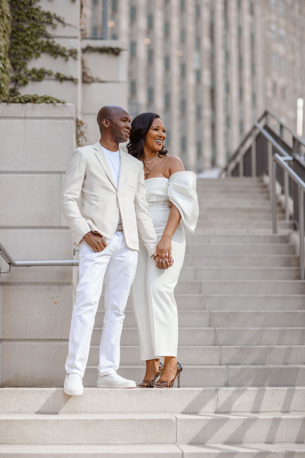 Elegant couple in matching white outfits pose together on city steps.