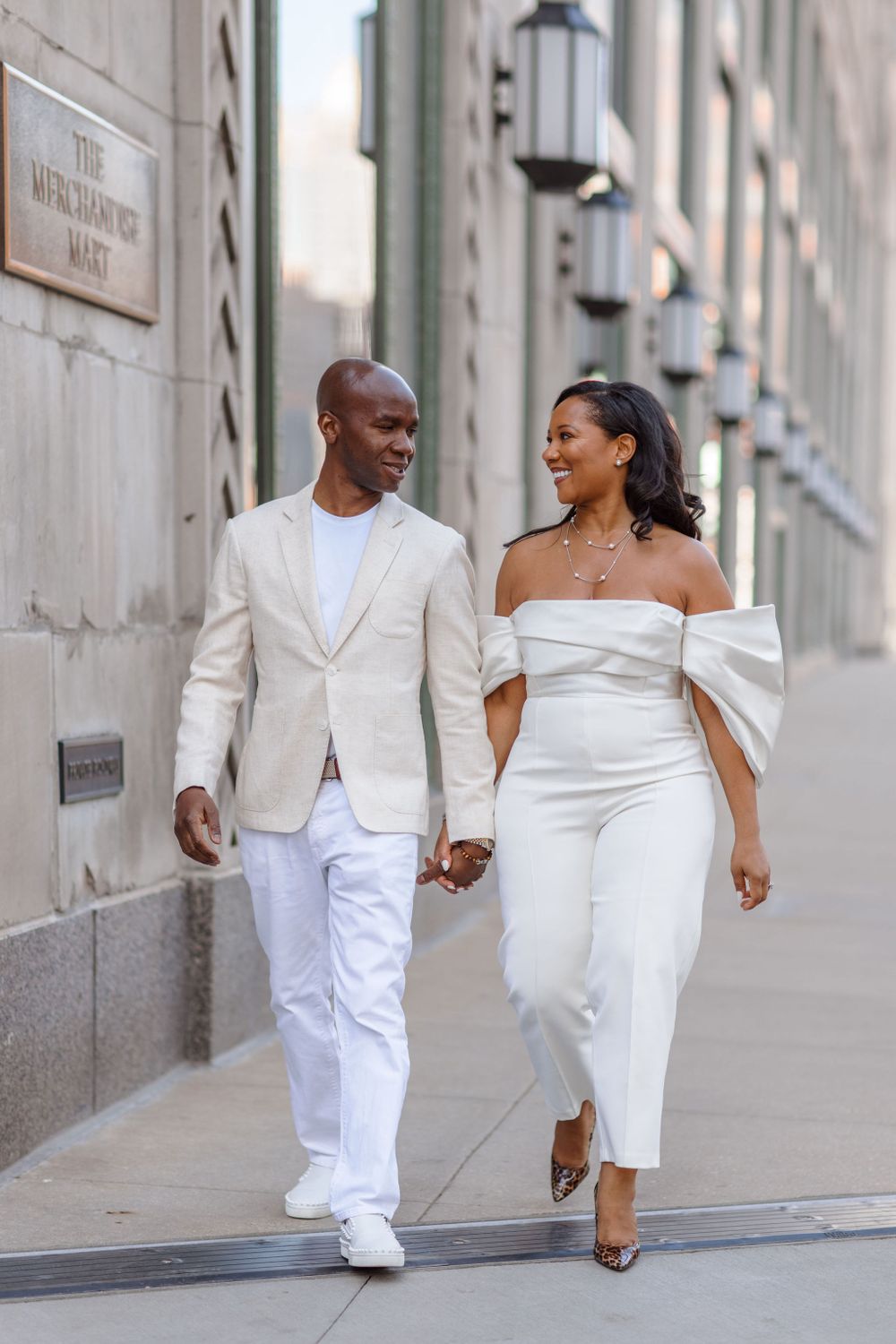 A couple in elegant all-white outfits walking hand in hand down a city street with lanterns in the background.