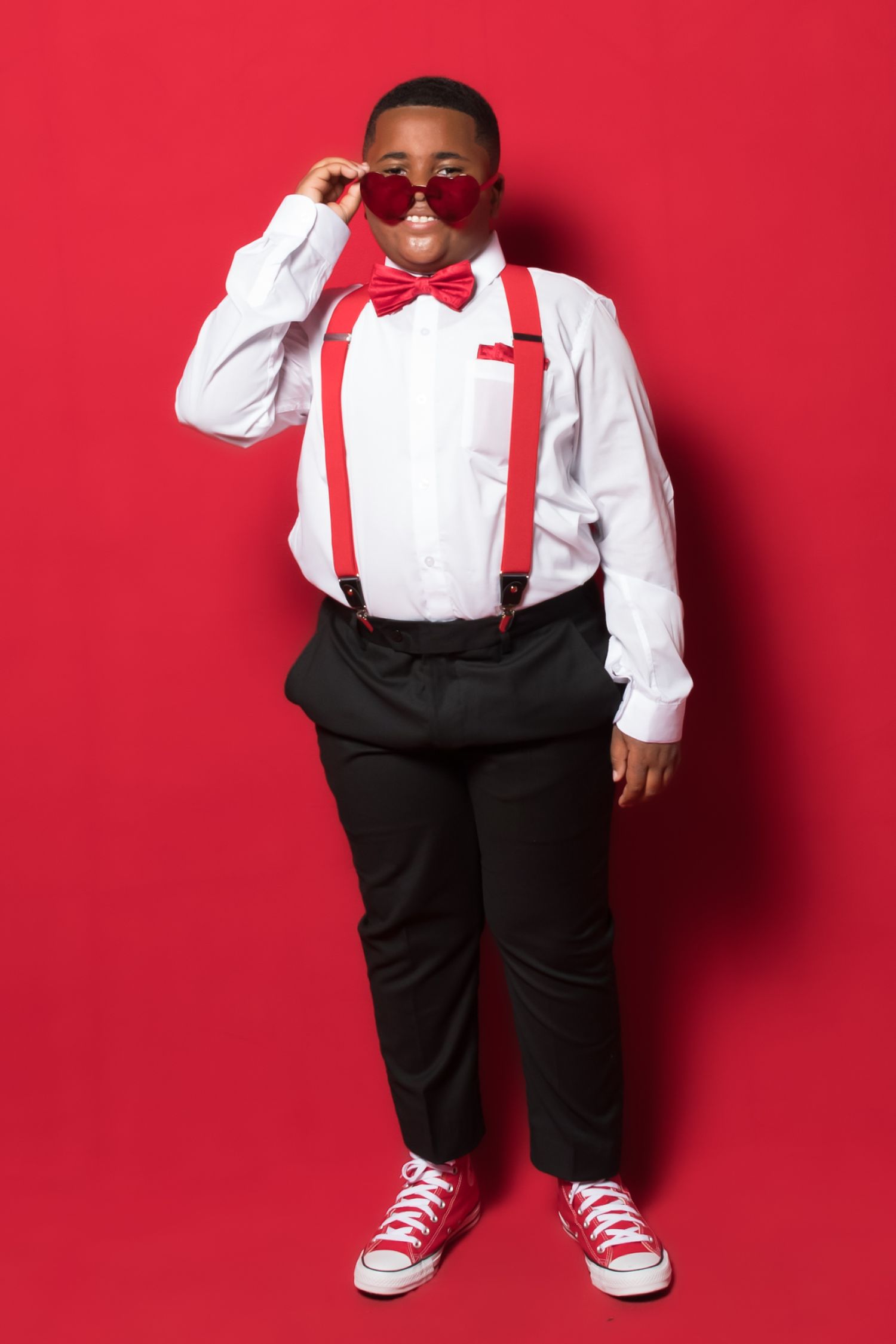 Stylish formal outfit featuring white dress shirt, red bow tie, black pants and red sneakers against red background.