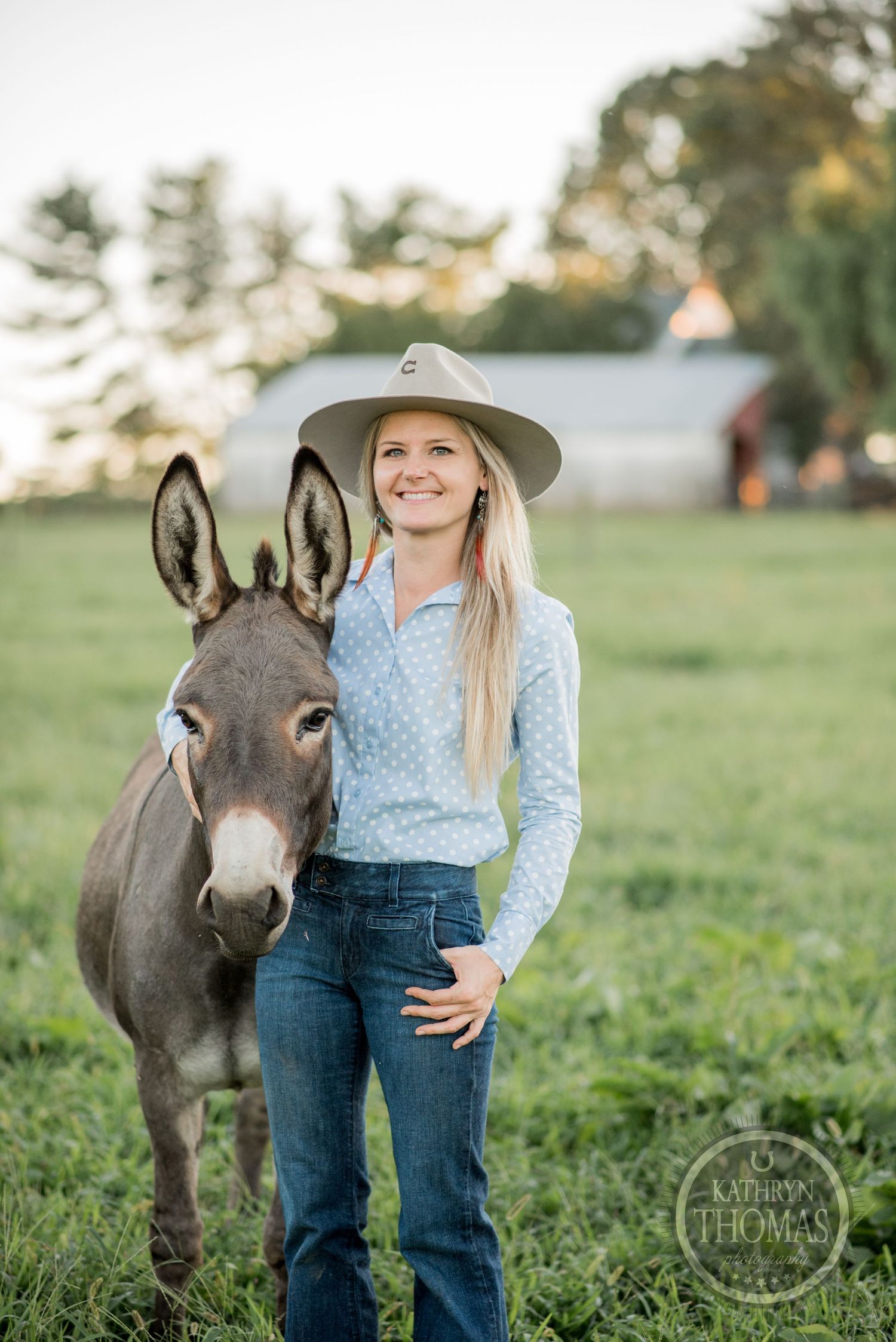 A friendly donkey stands next to someone in western wear in a green pasture.