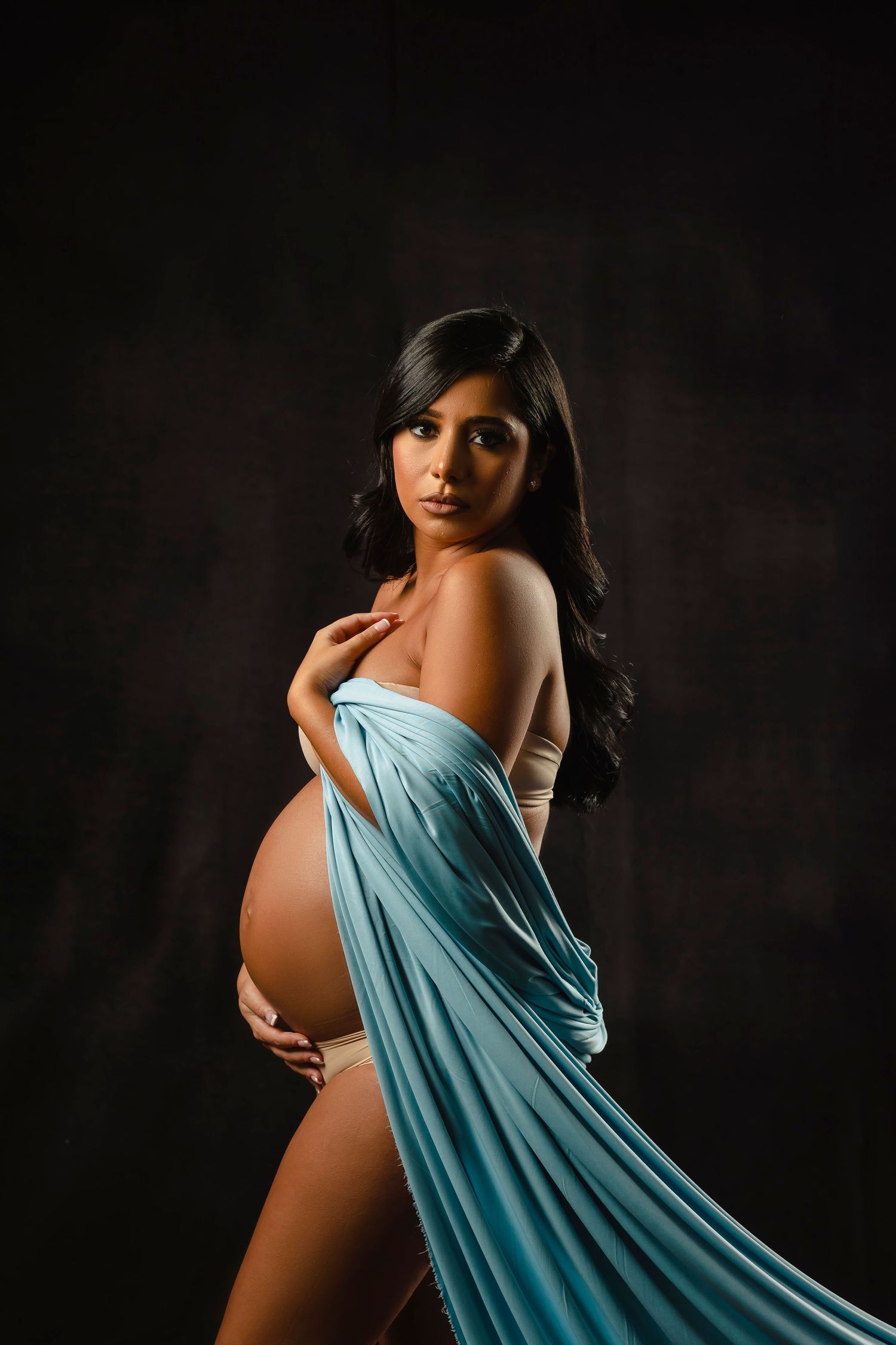 Artistic maternity photo series in turquoise fabric against dark background creates ethereal silhouette.