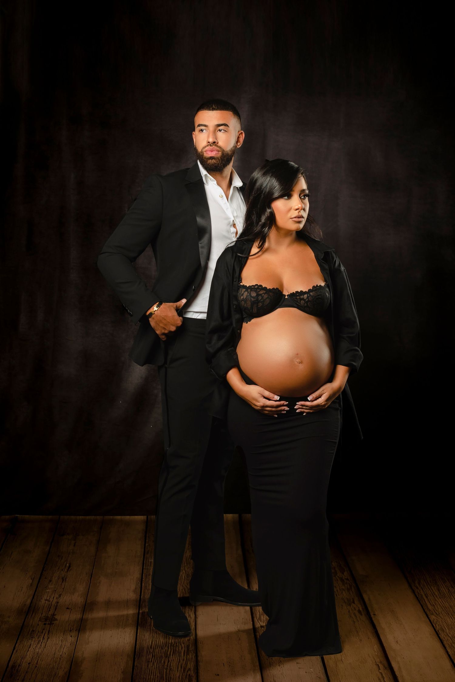Dramatic studio maternity session featuring couple in formal black attire on wooden floor.