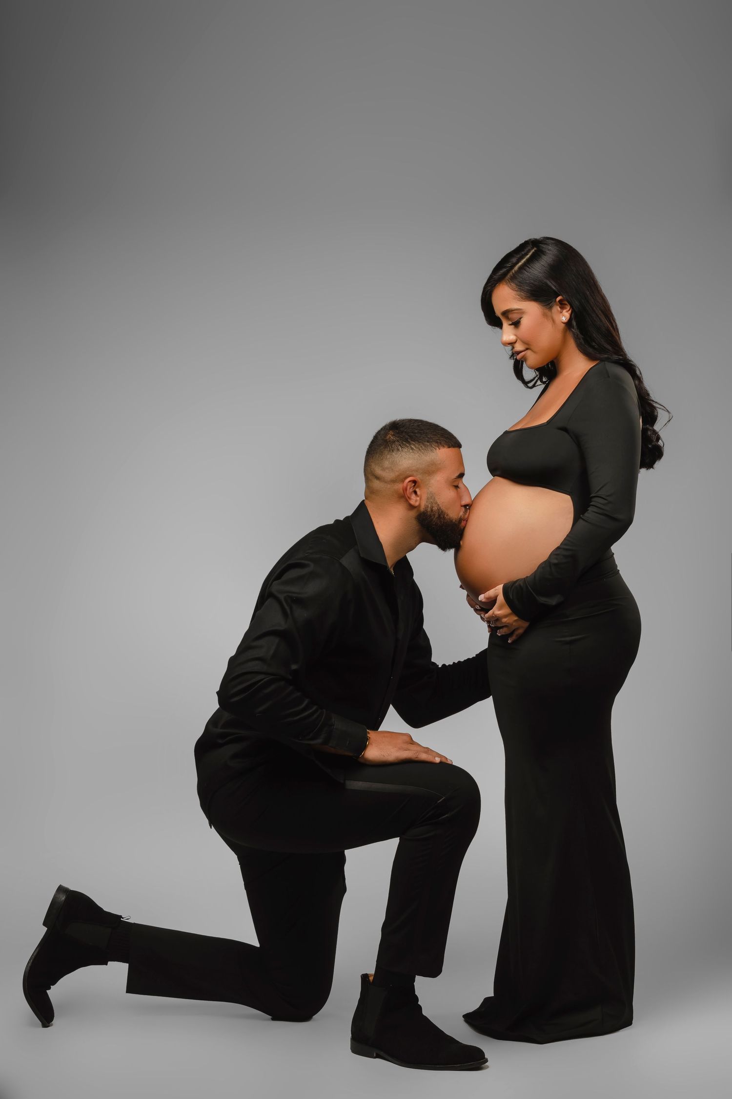 Intimate couple maternity photos in all black outfits against grey studio backdrop.