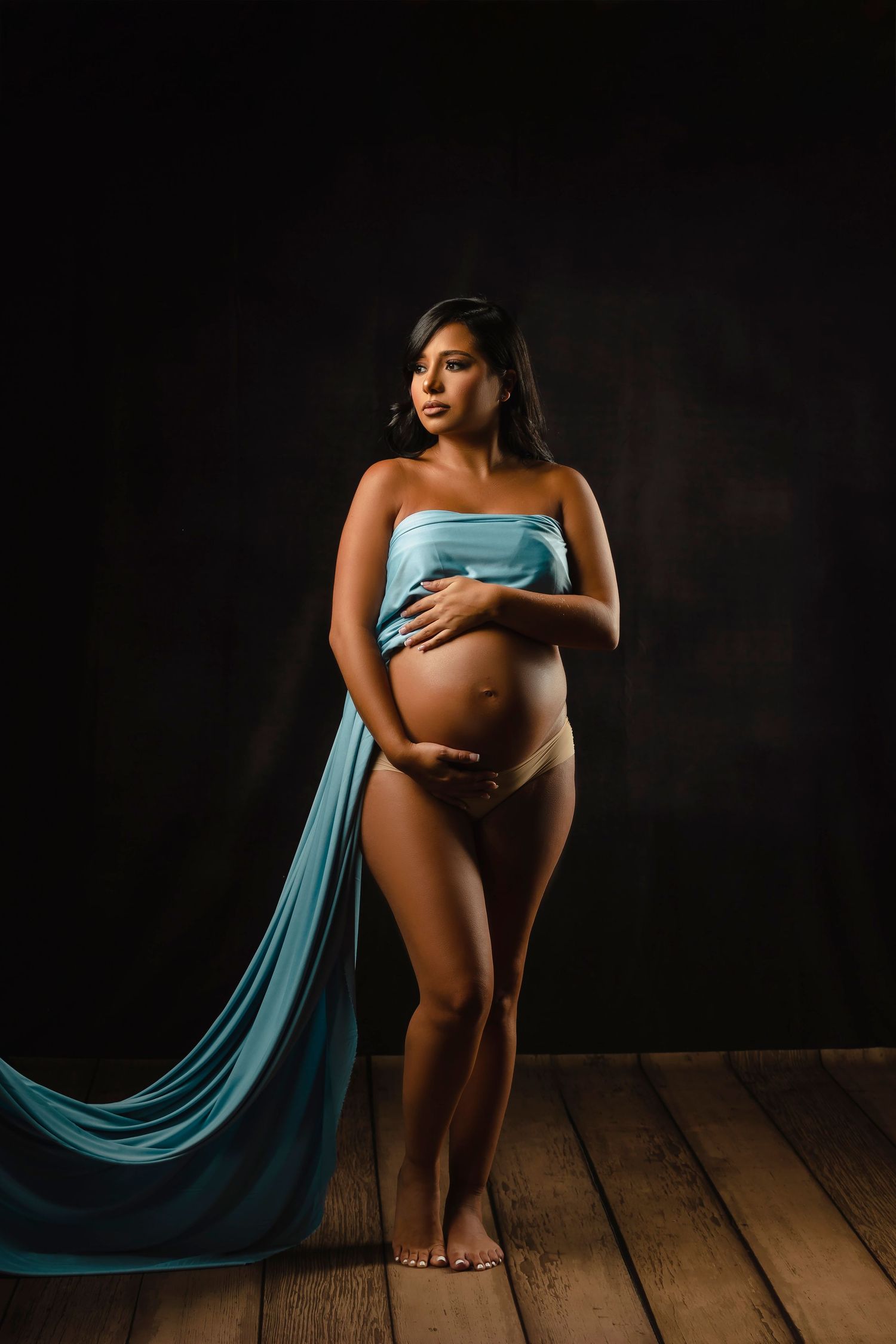 Artistic maternity portrait with flowing teal fabric in dramatic studio lighting.