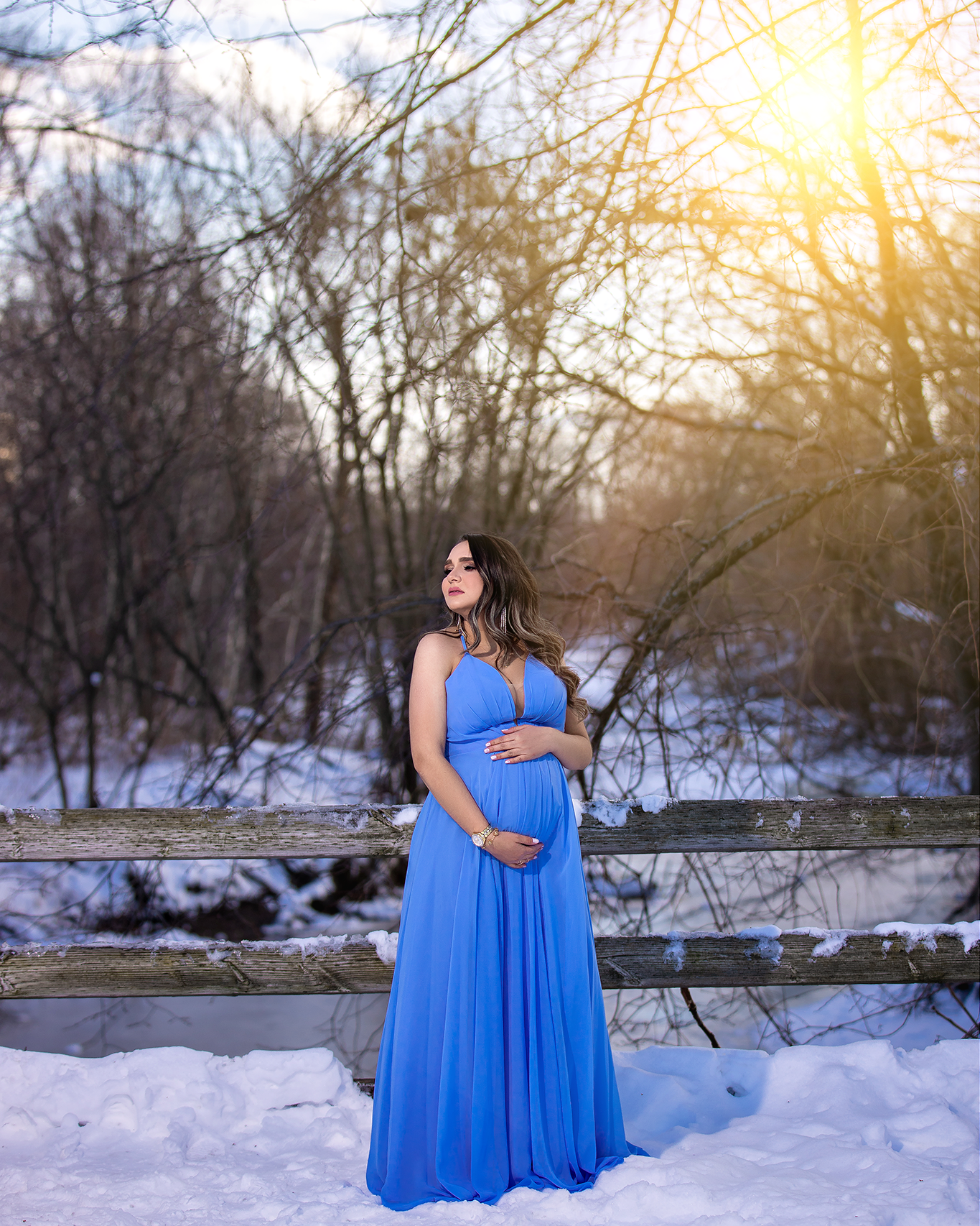 Maternity photo of woman in flowing blue dress standing in snowy winter landscape.
