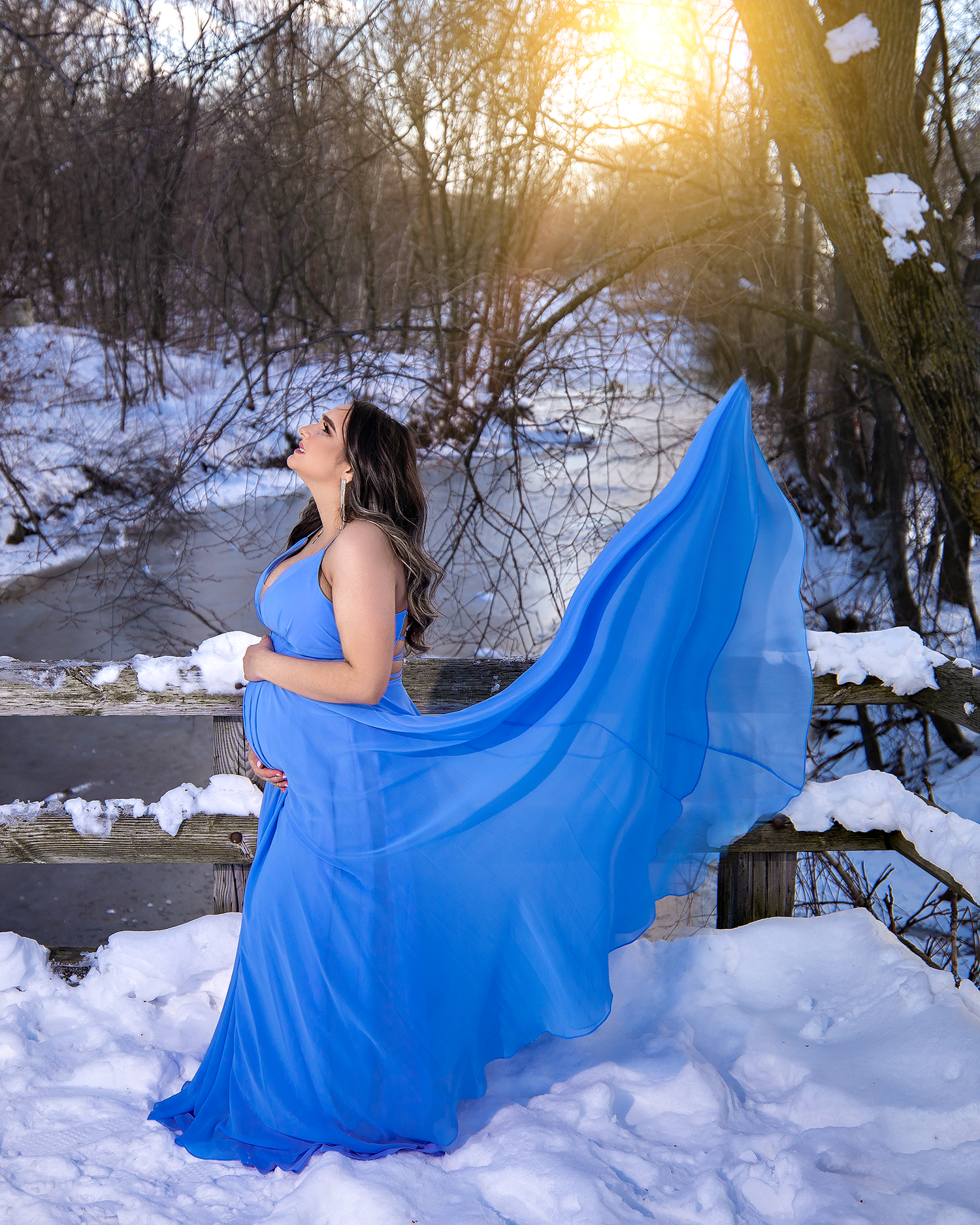 Graceful maternity portrait with flowing blue dress against snowy winter backdrop.