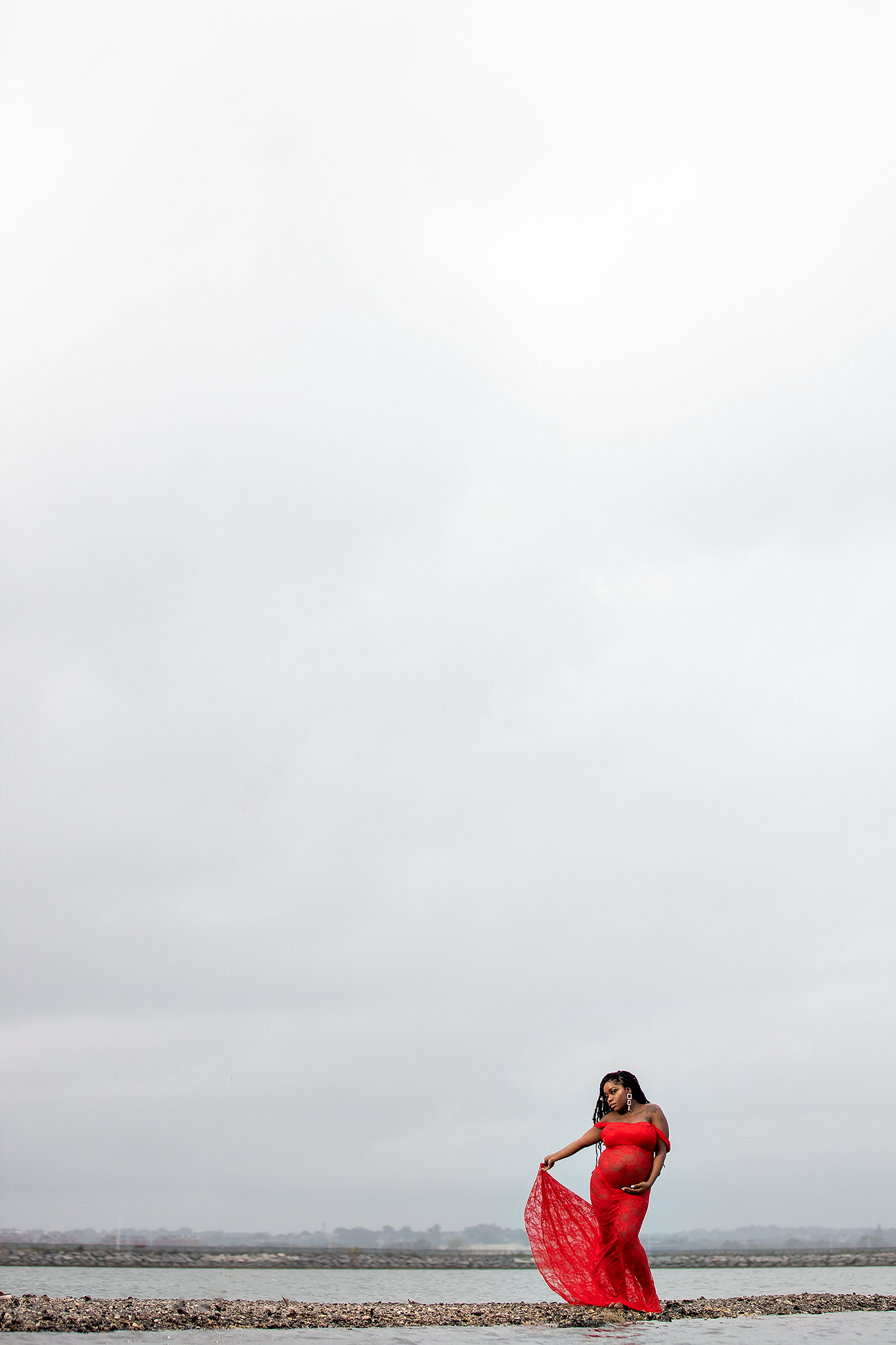 Minimalist maternity portrait with flowing red dress against grey sky.