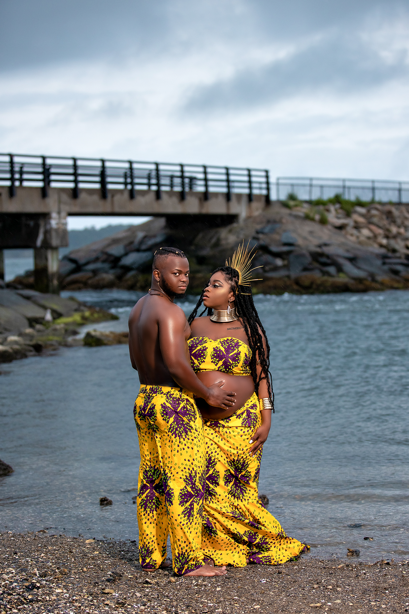Couple in matching yellow African print outfits pose for maternity photo by waterfront.