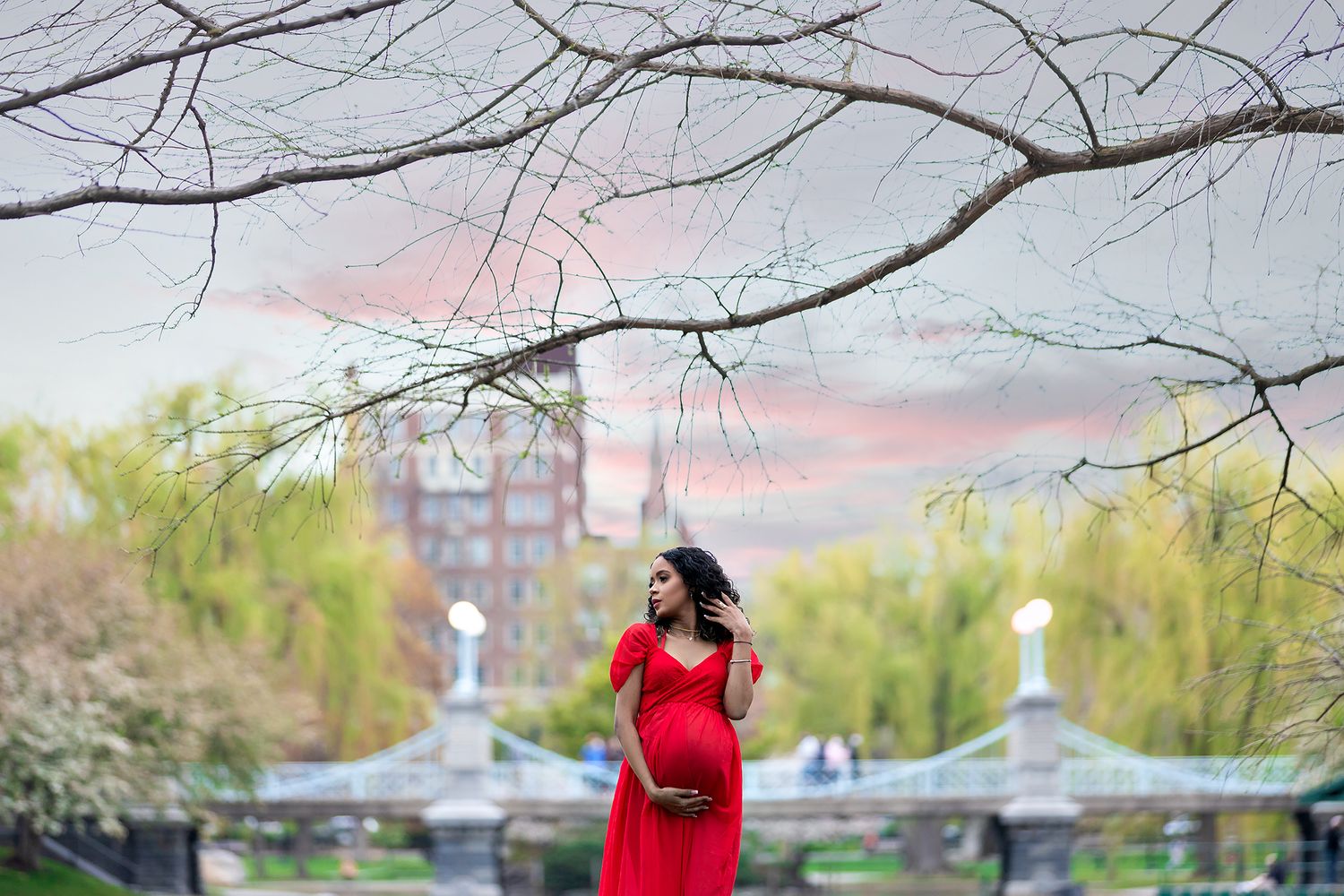 Maternity photo of person in red dress standing under bare branches at sunset.