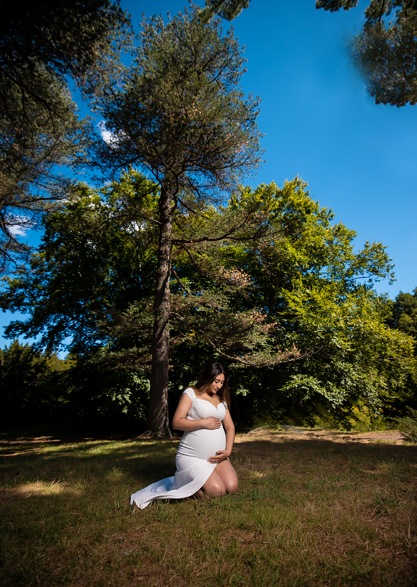 Person in white dress poses against tall pine trees in outdoor setting.