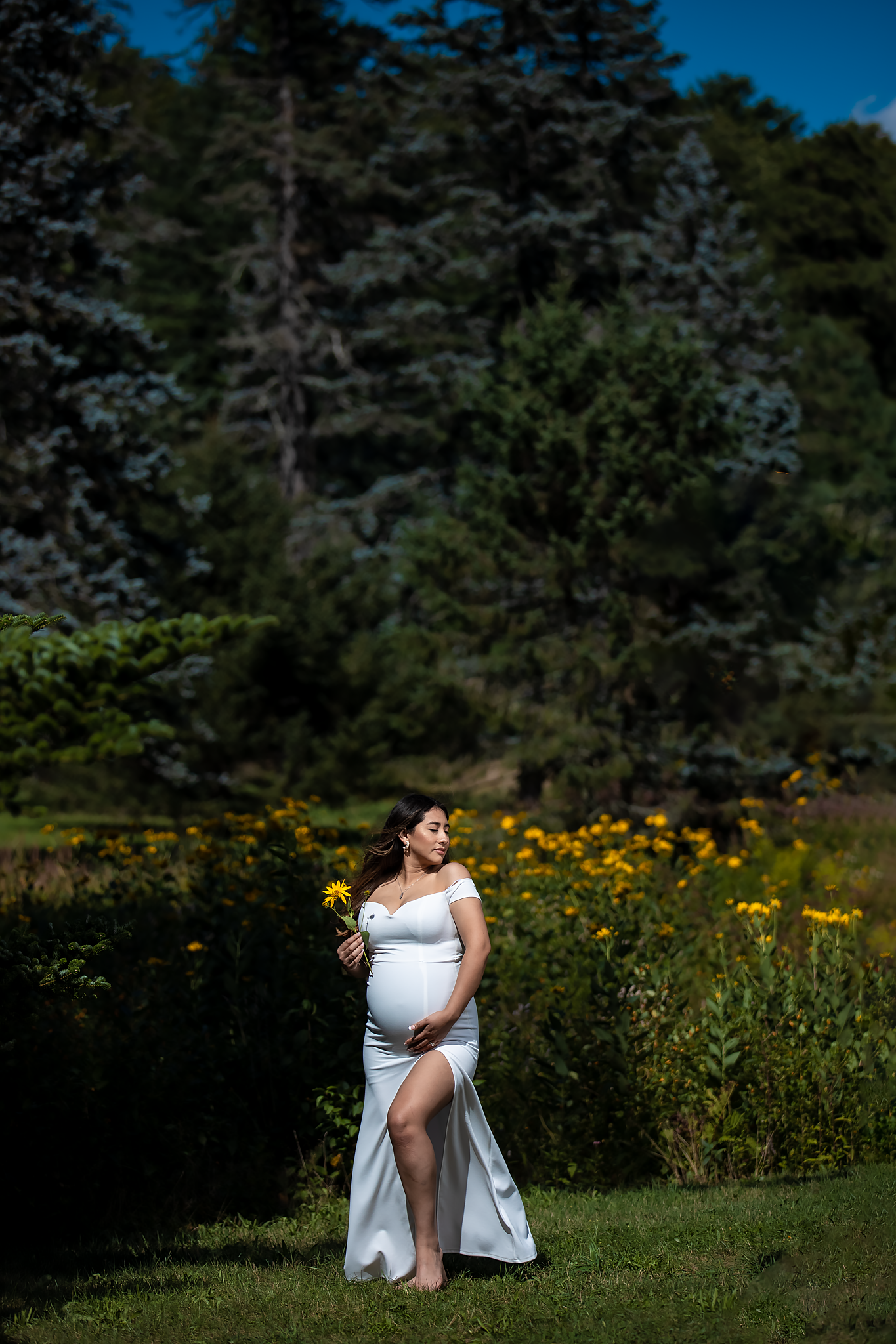 Person in white dress standing among wildflowers with evergreen forest backdrop.