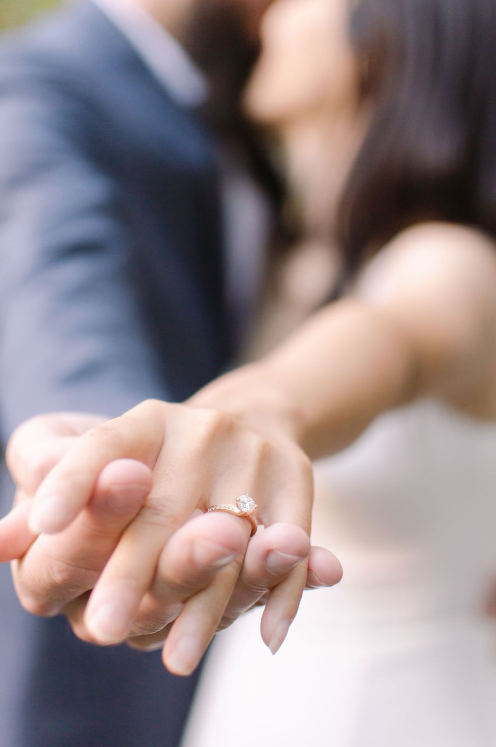 Close-up of an engagement ring on a womans finger as a couple shares a kiss in the background.