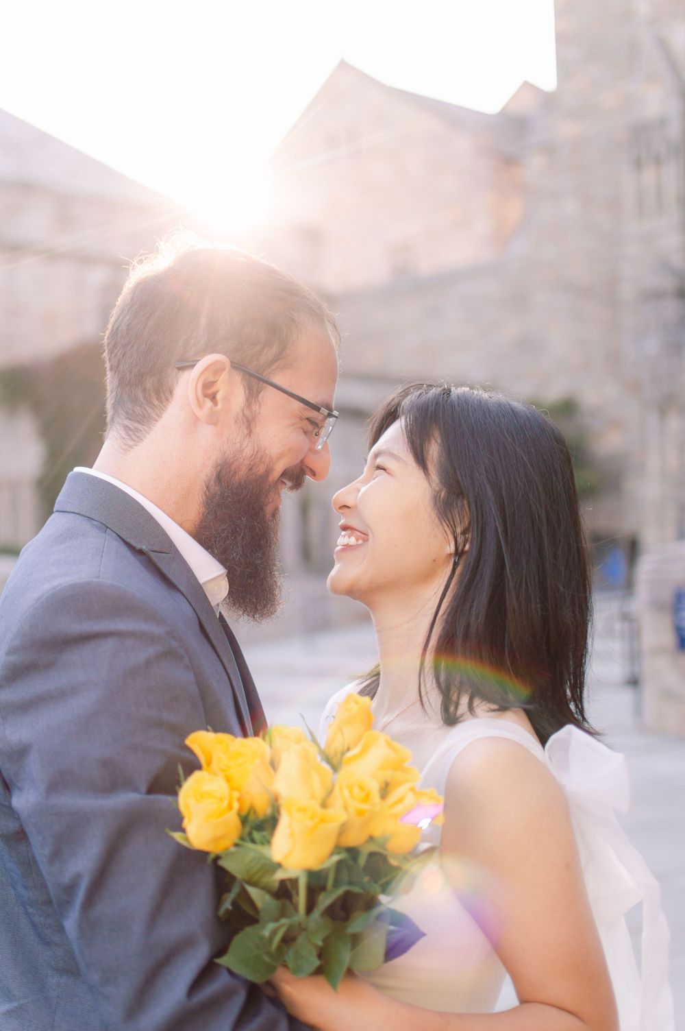 A joyful couple laughs together in golden sunlight, the bride holding a bouquet of bright yellow roses.