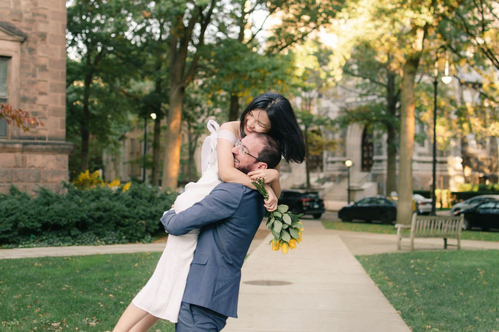 A groom lifts his bride in a joyful embrace on a tree-lined campus pathway during their wedding photos.
