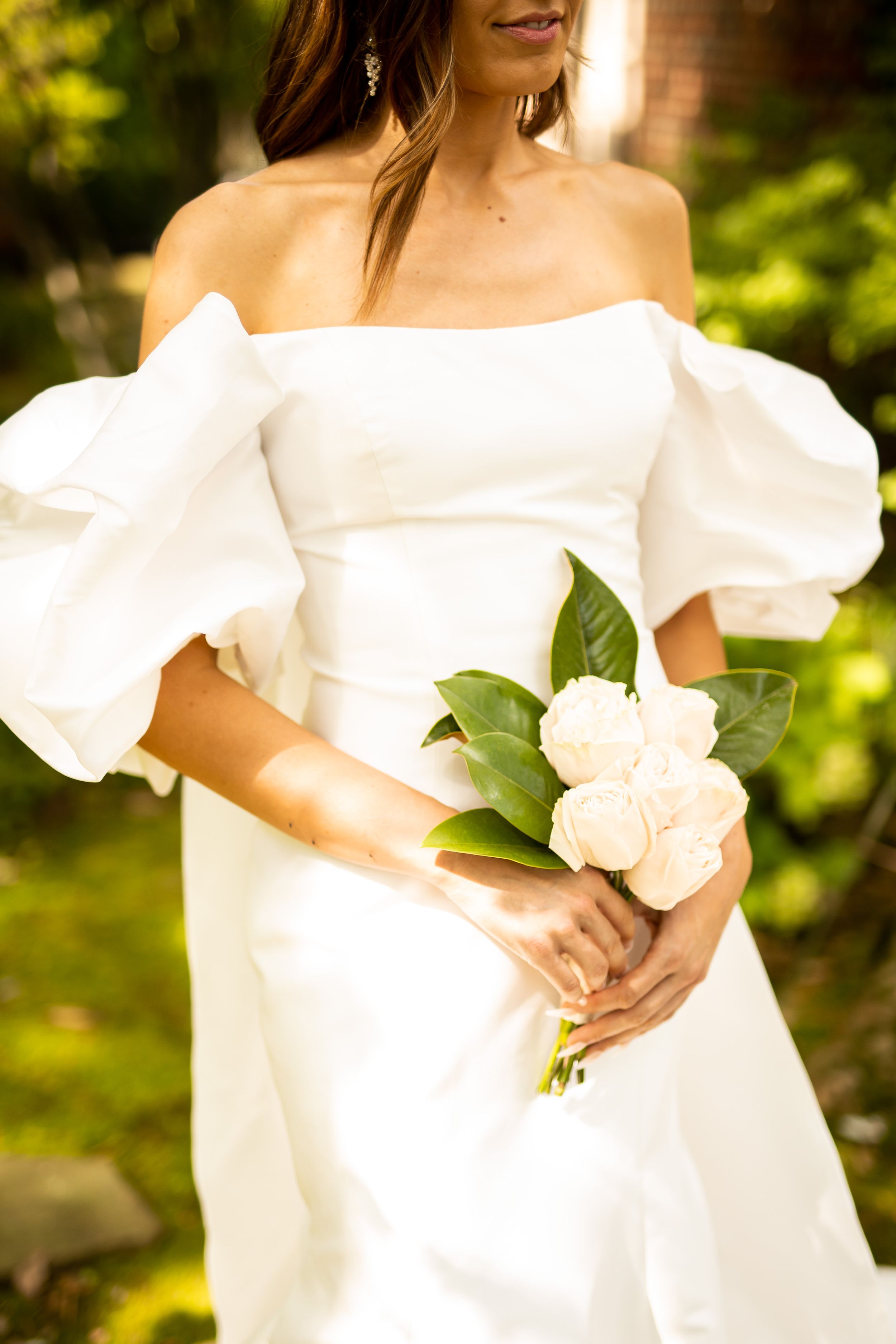 Bride in white dress holding a bouquet of pale roses against green foliage background.