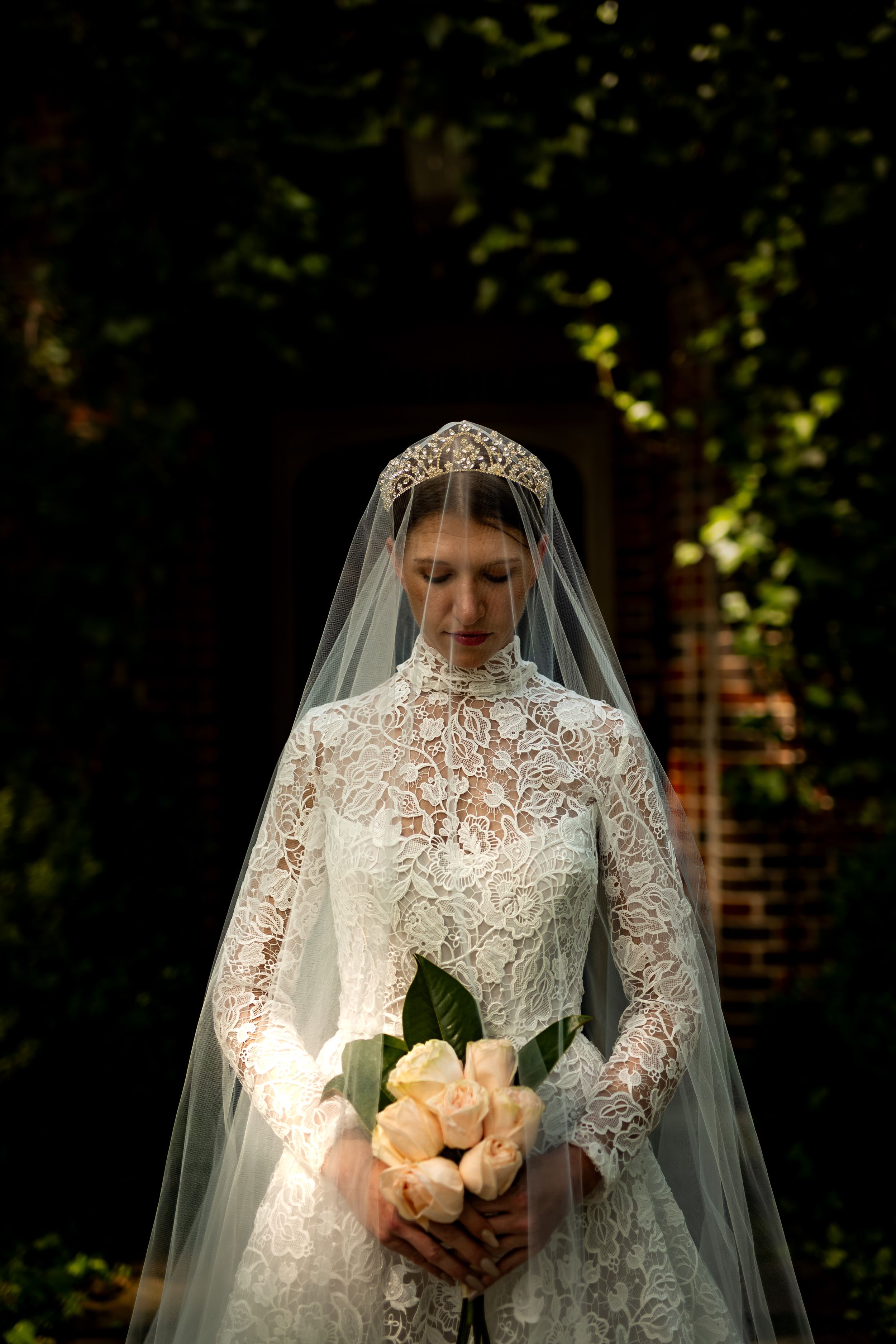 An elegant long-sleeved lace wedding gown with high neck detail and veil photographed in dramatic lighting.