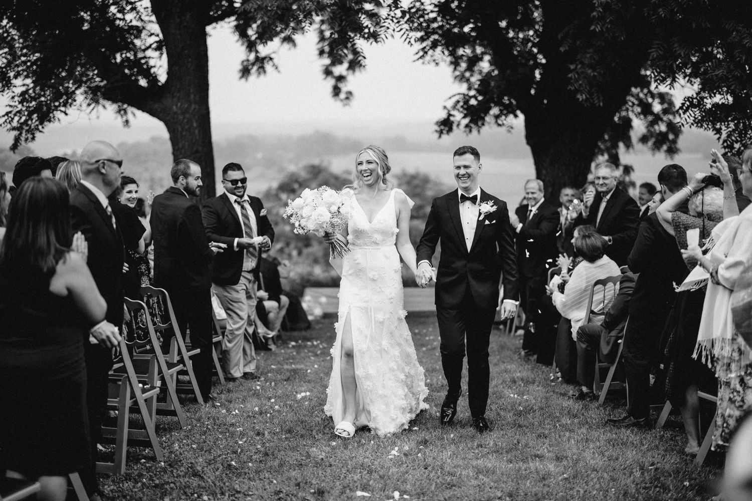 Black and white photo of outdoor wedding ceremony with guests seated in rows under trees.