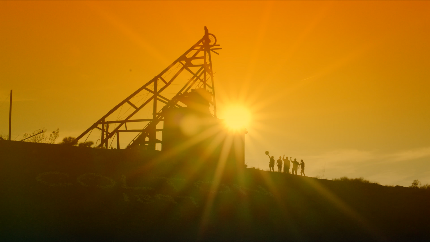 Silhouettes of people standing on a hill against a dramatic orange sunset with industrial equipment in view.