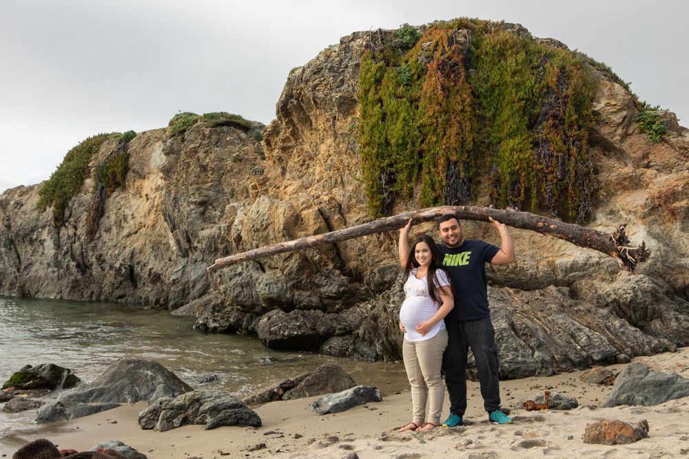 A couple poses in front of mossy coastal rocks and driftwood on a rugged beach.