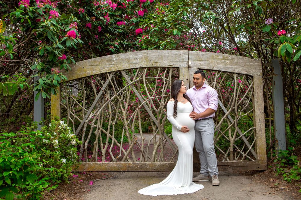 A couple poses by an ornate garden gate surrounded by flowering bushes and greenery.