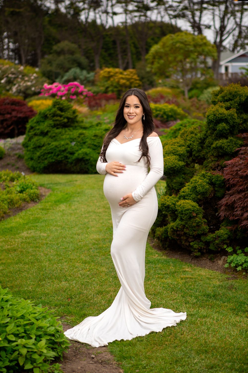 A maternity portrait in a flowing white dress amid colorful garden landscaping.