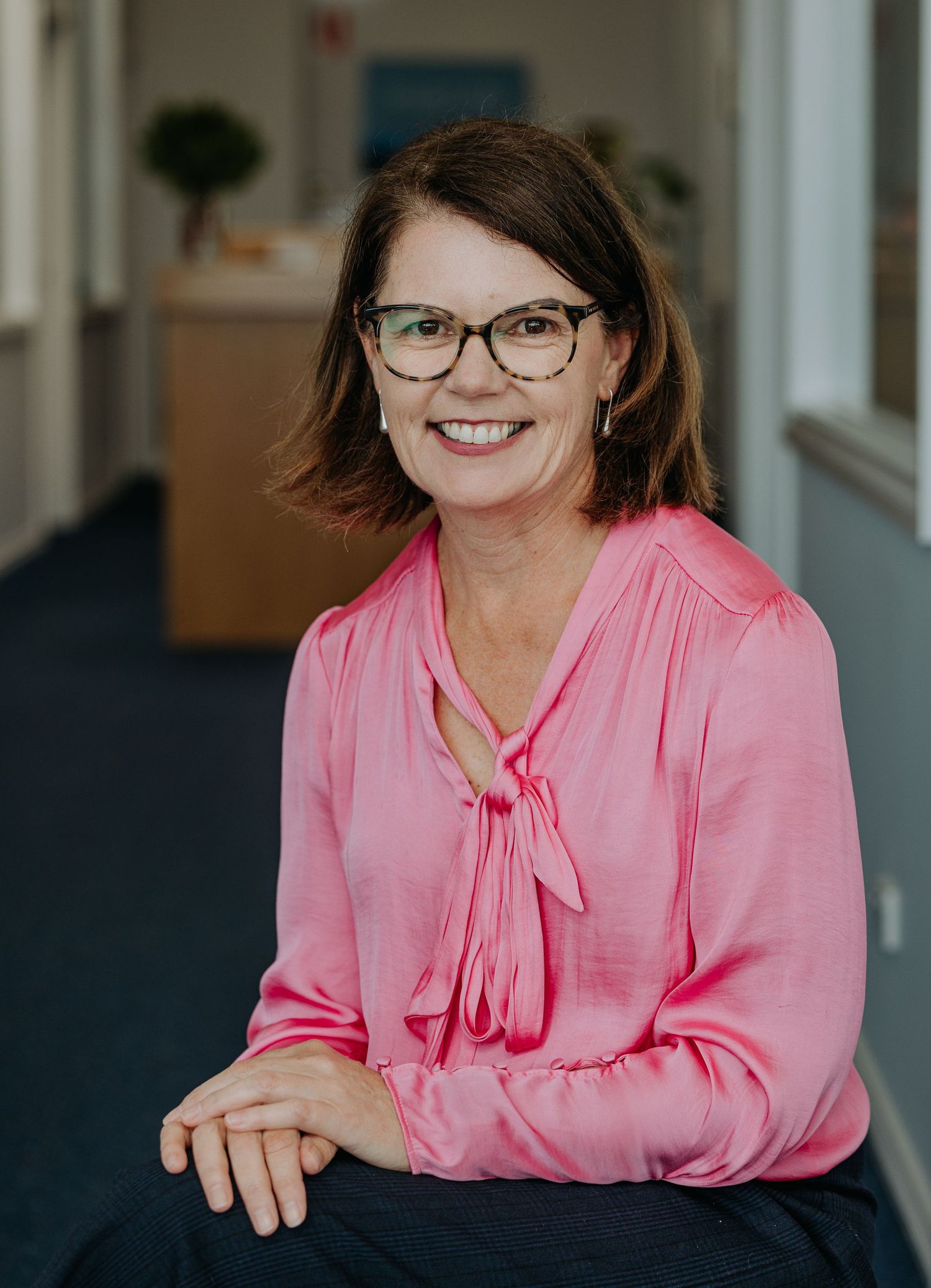 Professional headshot of someone wearing glasses and a pink blouse with a bow tie neckline in an office setting.