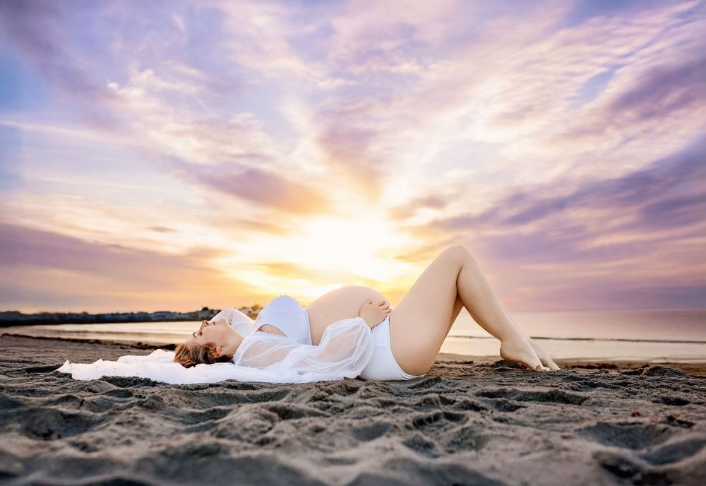 Maternity photo of an expectant mother in a white gown lying on the sand  New Hampshire pregnancy sunrise maternity photographer