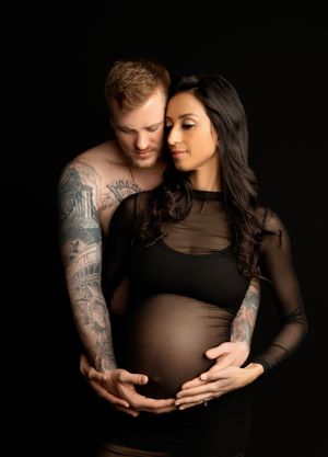 Studio maternity photo of a couple holding the baby bump, wearing black outfits against a dark background. Stephanie Ann Photo New Hampshire