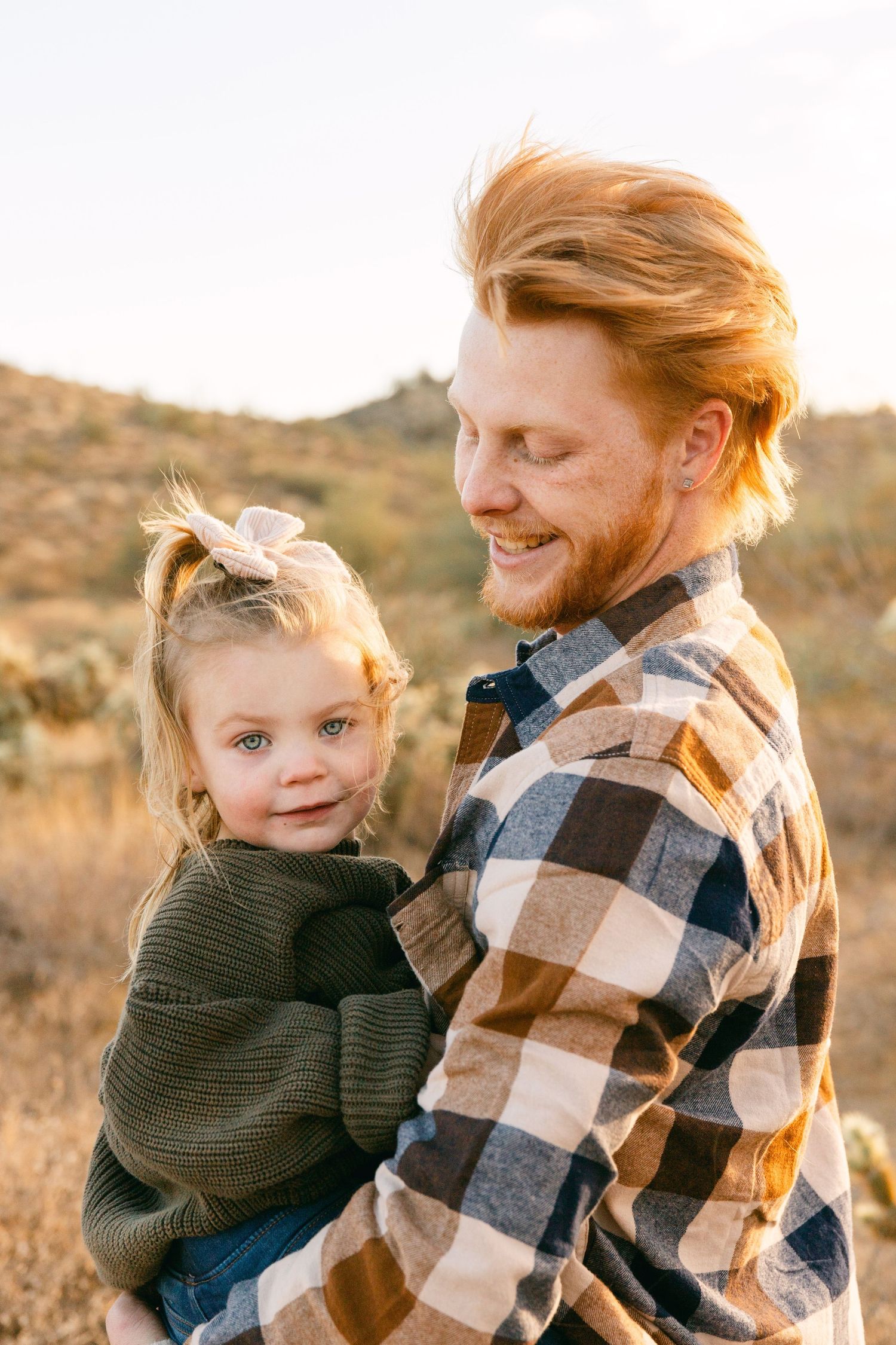 A man in a flannel shirt is smiling at the toddler he's holding. She has her hair pulled back and is wearing a green sweater.