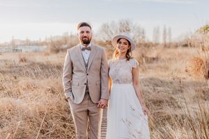 Ein Paar in Hochzeitskleidung steht auf einem Holzweg inmitten von hohem trockenem Gras.