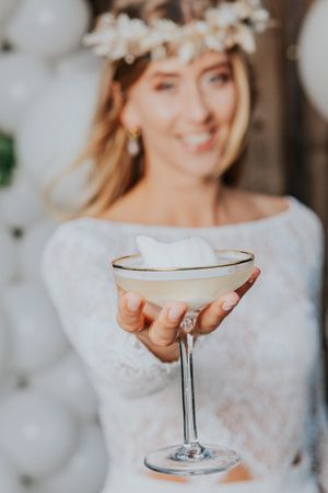 Nahaufnahme eines Champagnerglases, das von einer Person gehalten wird, die ein weißes Spitzenkleid und eine Blumenkrone trägt.