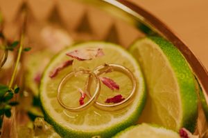 Die Eheringe ruhen zart auf einer frischen Limettenscheibe, die mit rosa Blütenblättern garniert ist.
