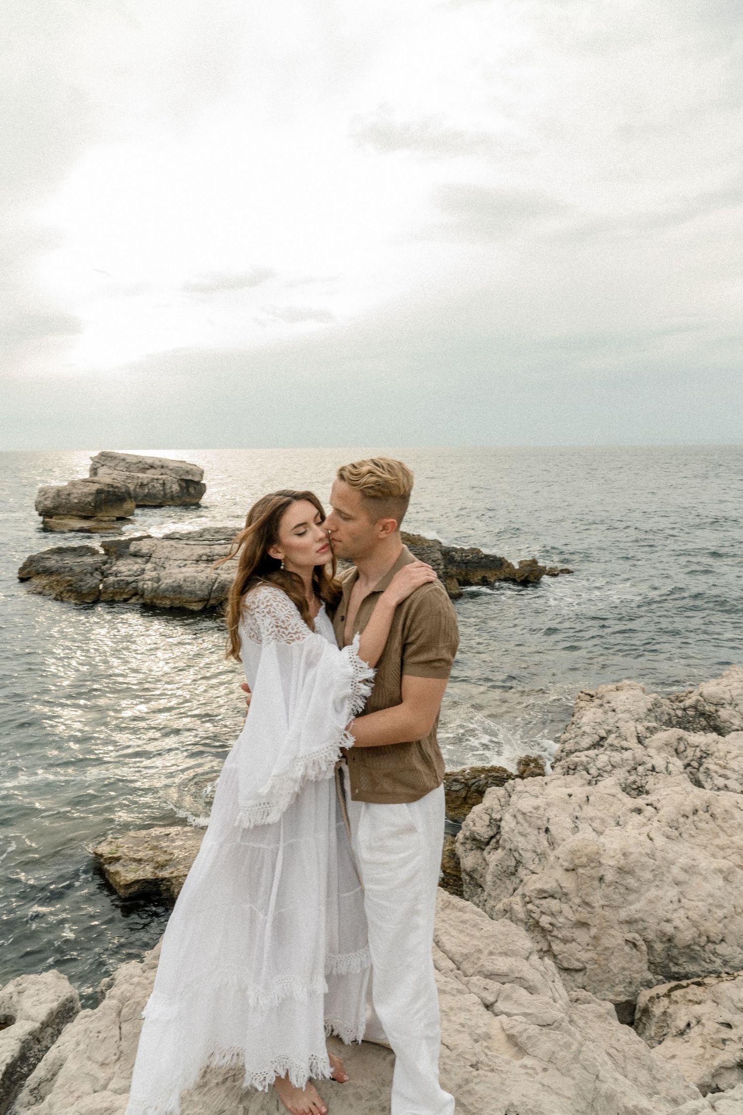 A couple in white embraces on rocky coastal cliffs against a misty ocean backdrop.