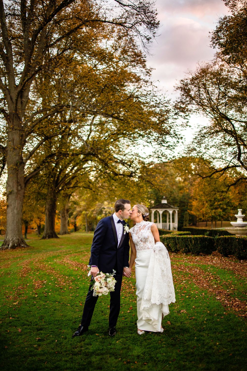 A couple shares an intimate moment under autumn trees near a gazebo in a picturesque garden setting.