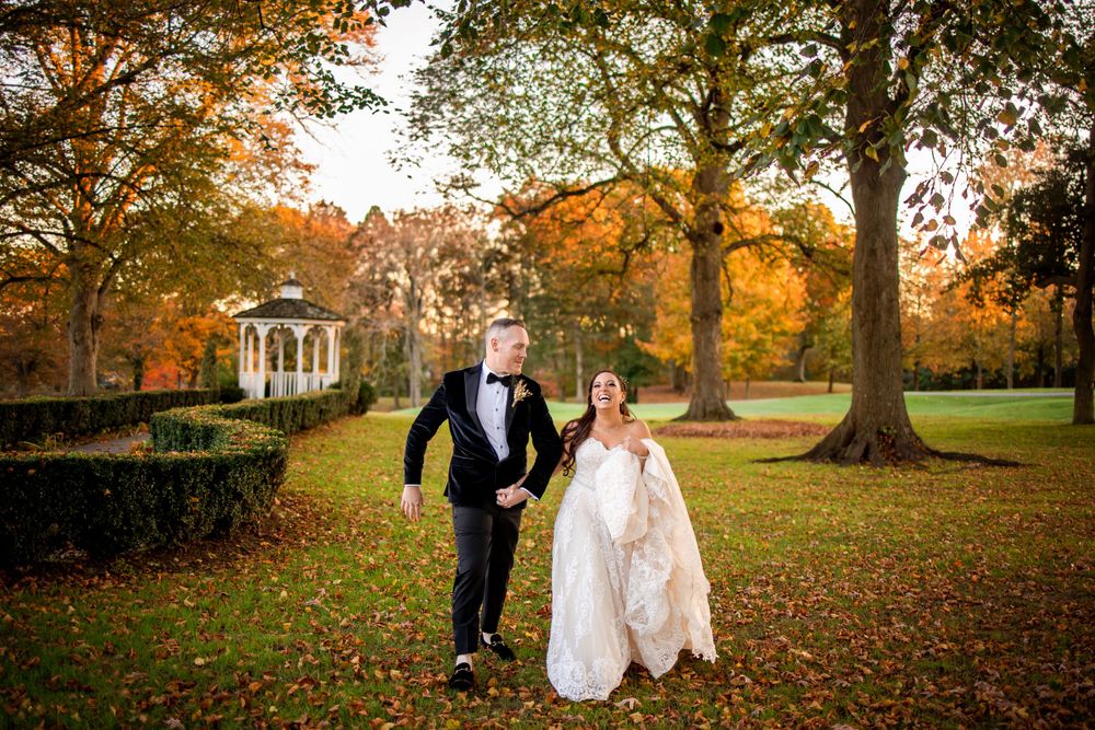Couple walks hand in hand through fall foliage with white gazebo in background.