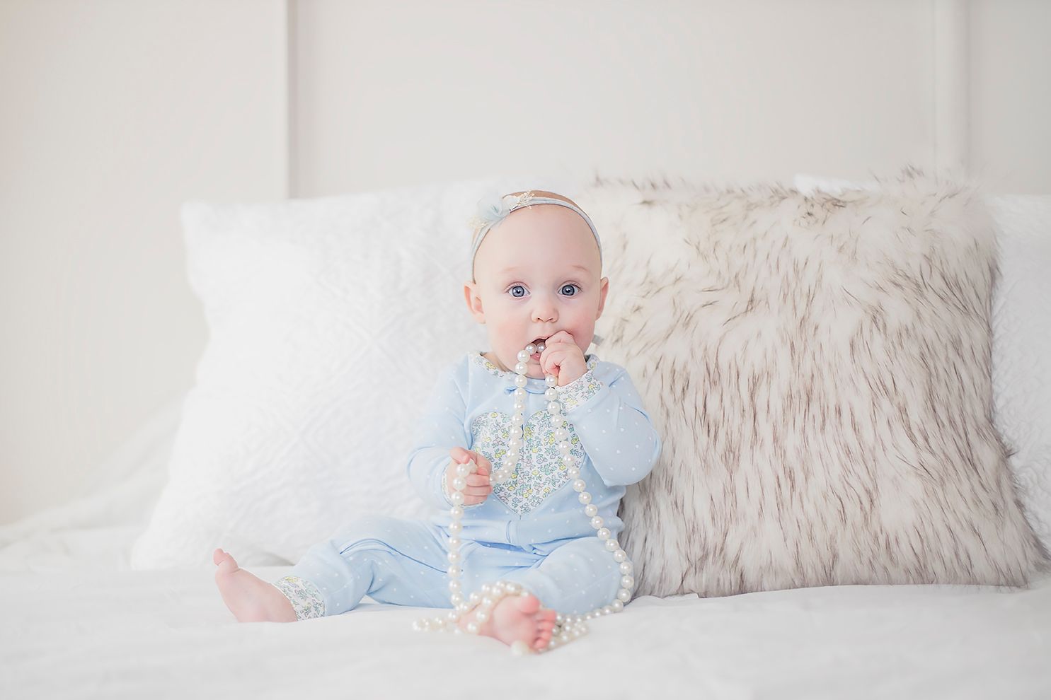 Baby in light blue outfit sitting on bed with plush pillows.