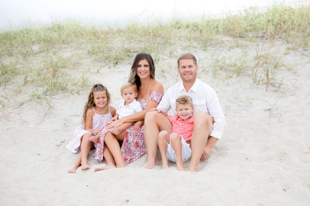 A family group sits together on sandy beach dunes wearing coordinated light summer outfits.