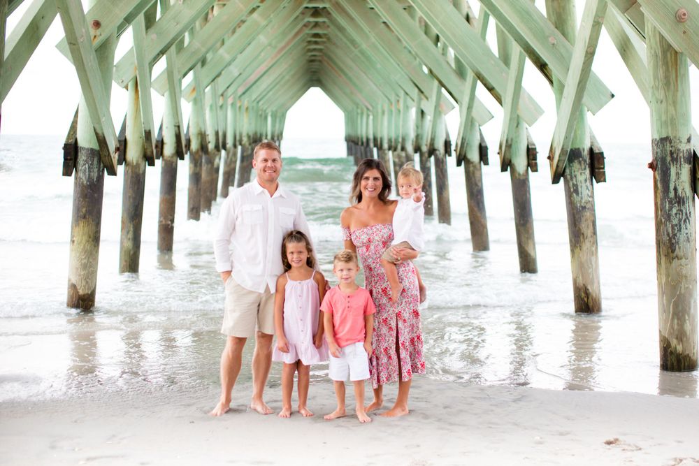 A family in coordinated pastel outfits poses under a wooden pier at the beach.