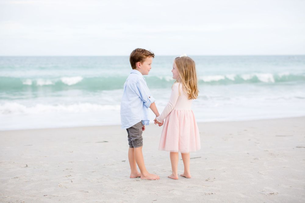 Two children holding hands while standing barefoot on a sandy beach.