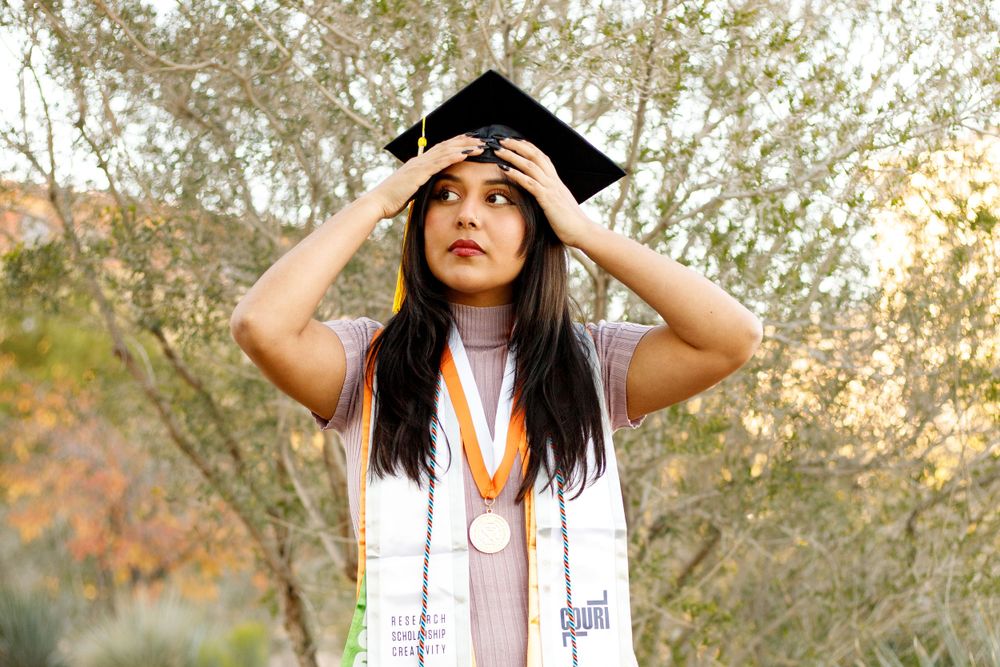 Female graduate adjusting her cap while wearing graduation regalia and medals outdoors.