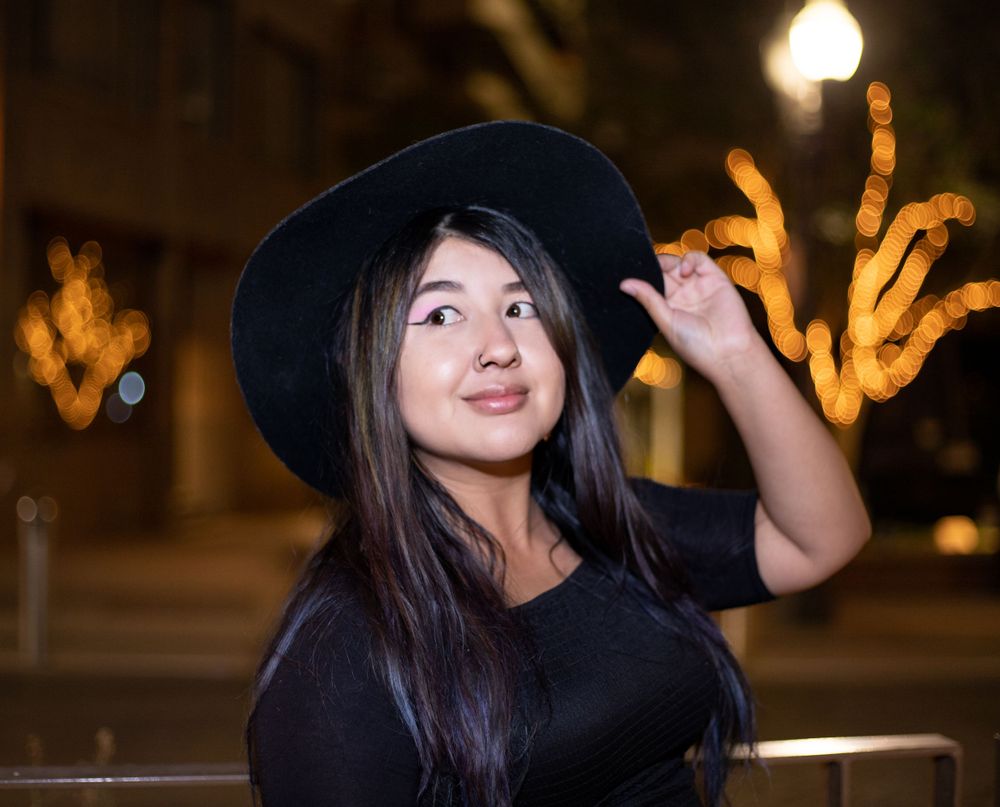 Young Asian woman in black outfit and wide-brim hat posing at night with golden bokeh lights behind her.
