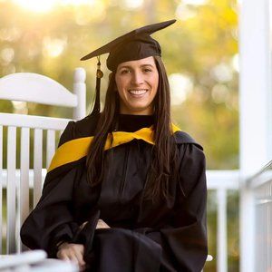 Someone in graduation regalia poses on a white porch bench at sunset.